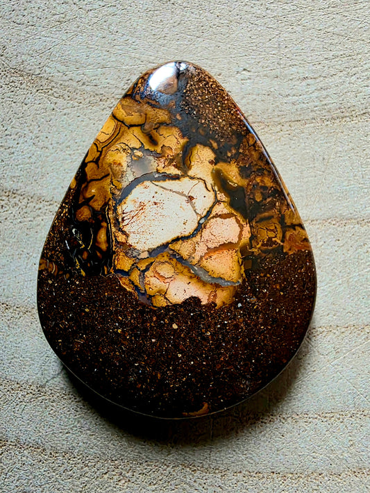 A polished teardrop-shaped stone with a mottled pattern of dark brown, golden yellow, and cream, resting on a textured light grey surface.