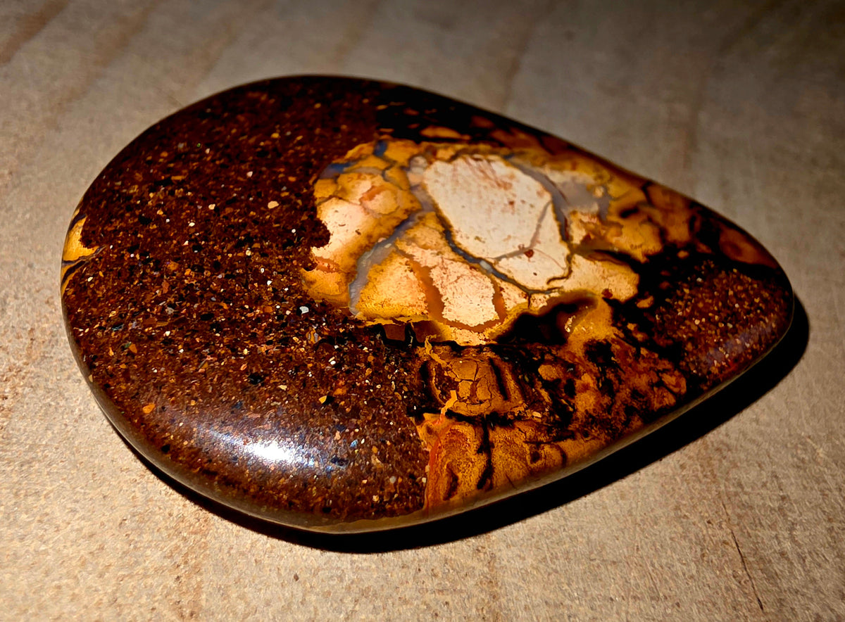 A polished brown and gold boulder stone with a central cream-coloured matrix, photographed on a textured beige surface.