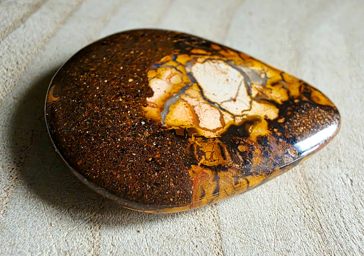 A polished teardrop-shaped stone with a mottled pattern of dark brown, golden yellow, and cream, resting on a textured beige background.