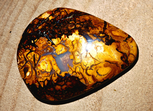 A polished natural stone cabochon with a marbled pattern of golden yellow, amber, and black, resting on a textured beige surface.
