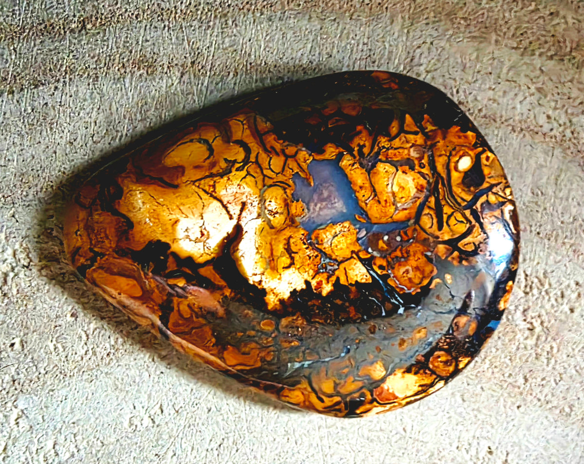 A polished teardrop-shaped cabochon with a marbled pattern of metallic gold, orange, black, and blue, resting on a textured beige background.