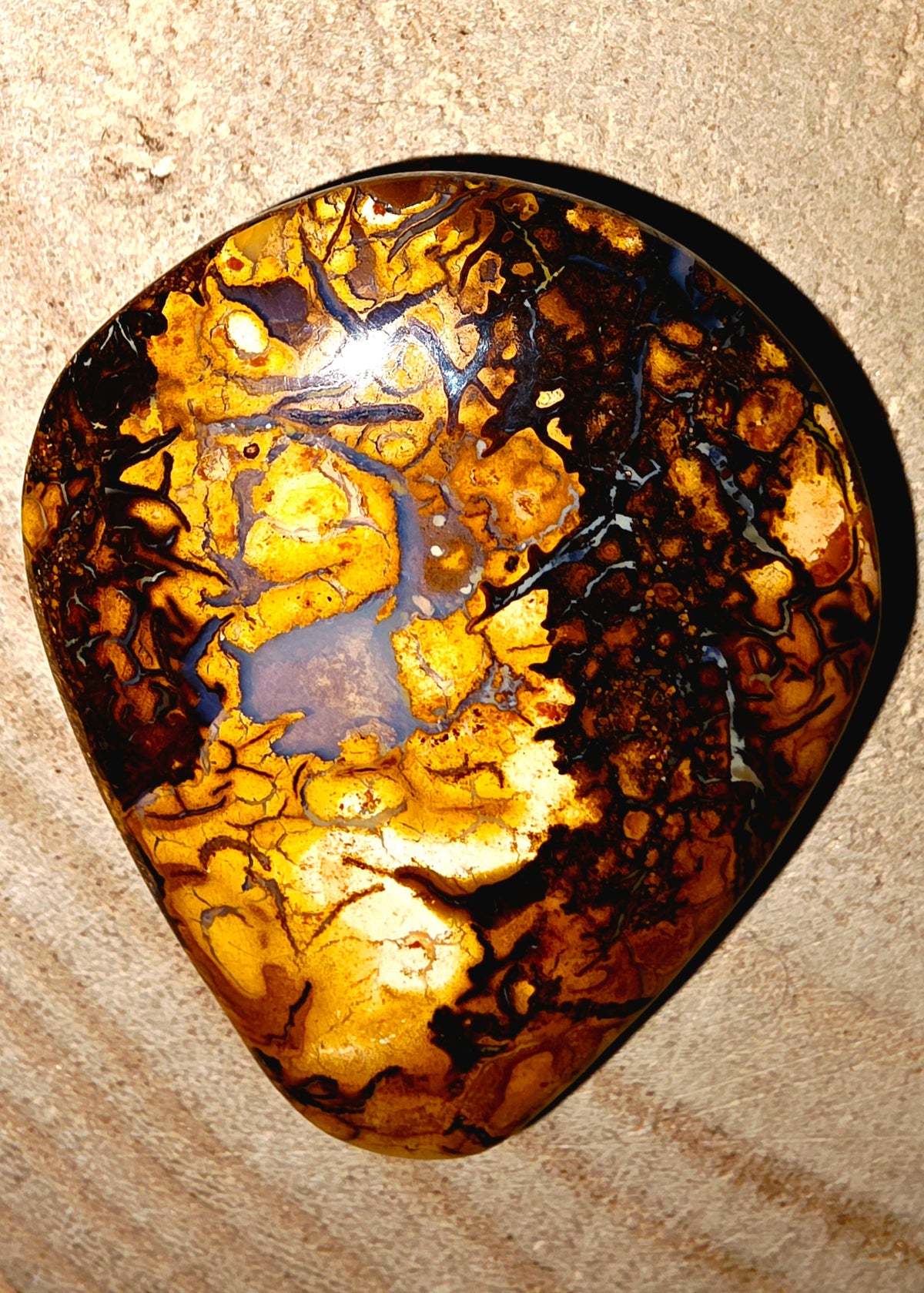 A polished teardrop-shaped gemstone with a marbled pattern of golden yellow, amber, and dark brown, resting on a sandy beige background.