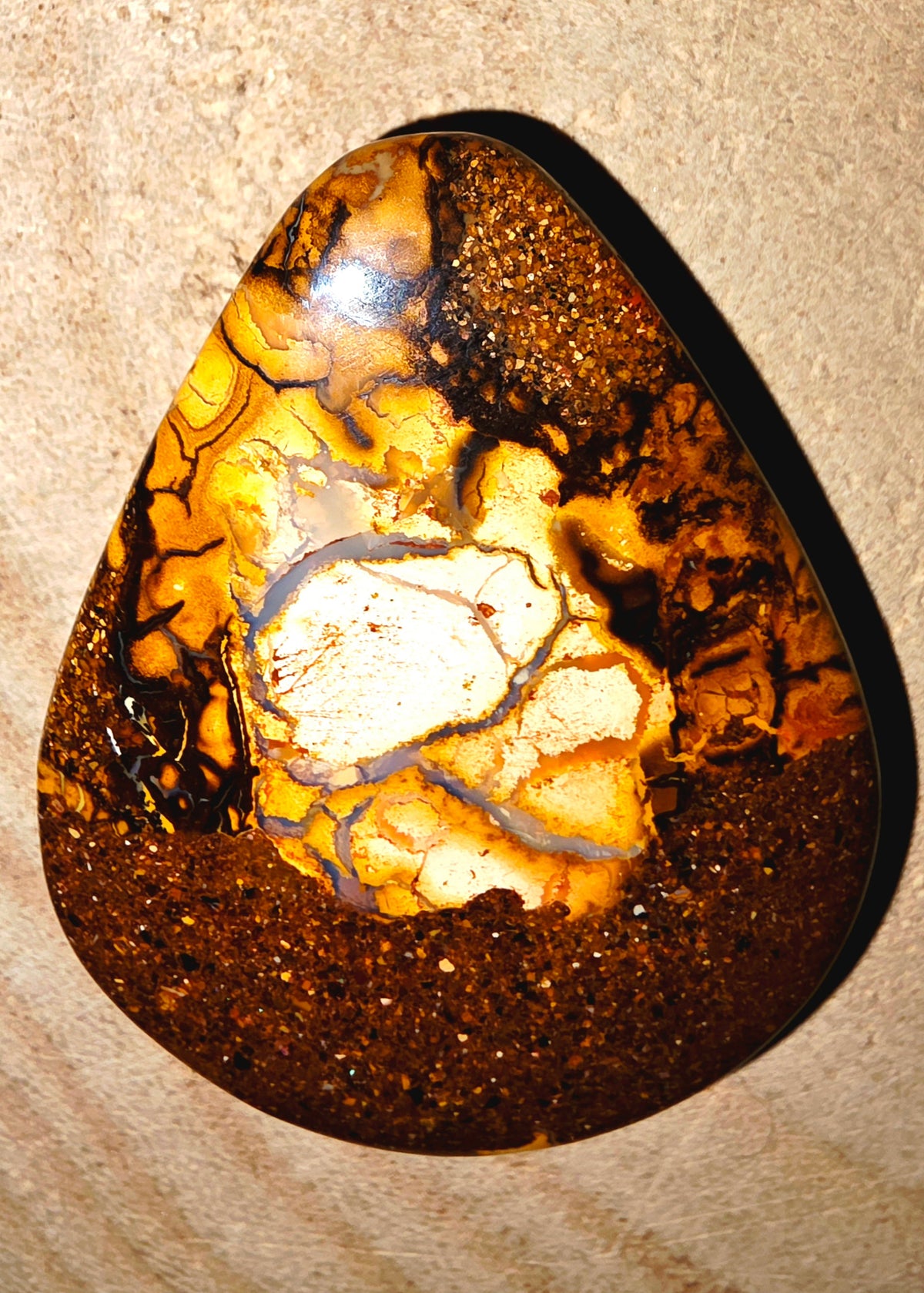 A polished teardrop-shaped gemstone with a rich pattern of golden yellow, deep brown, and black matrix, photographed close-up against a textured beige background.