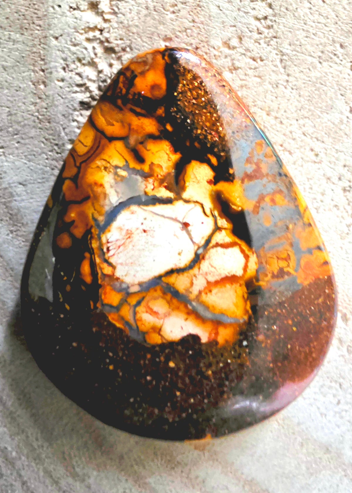 A polished teardrop-shaped stone with a marbled pattern of black, orange, brown, and cream, resting on a textured light grey surface.