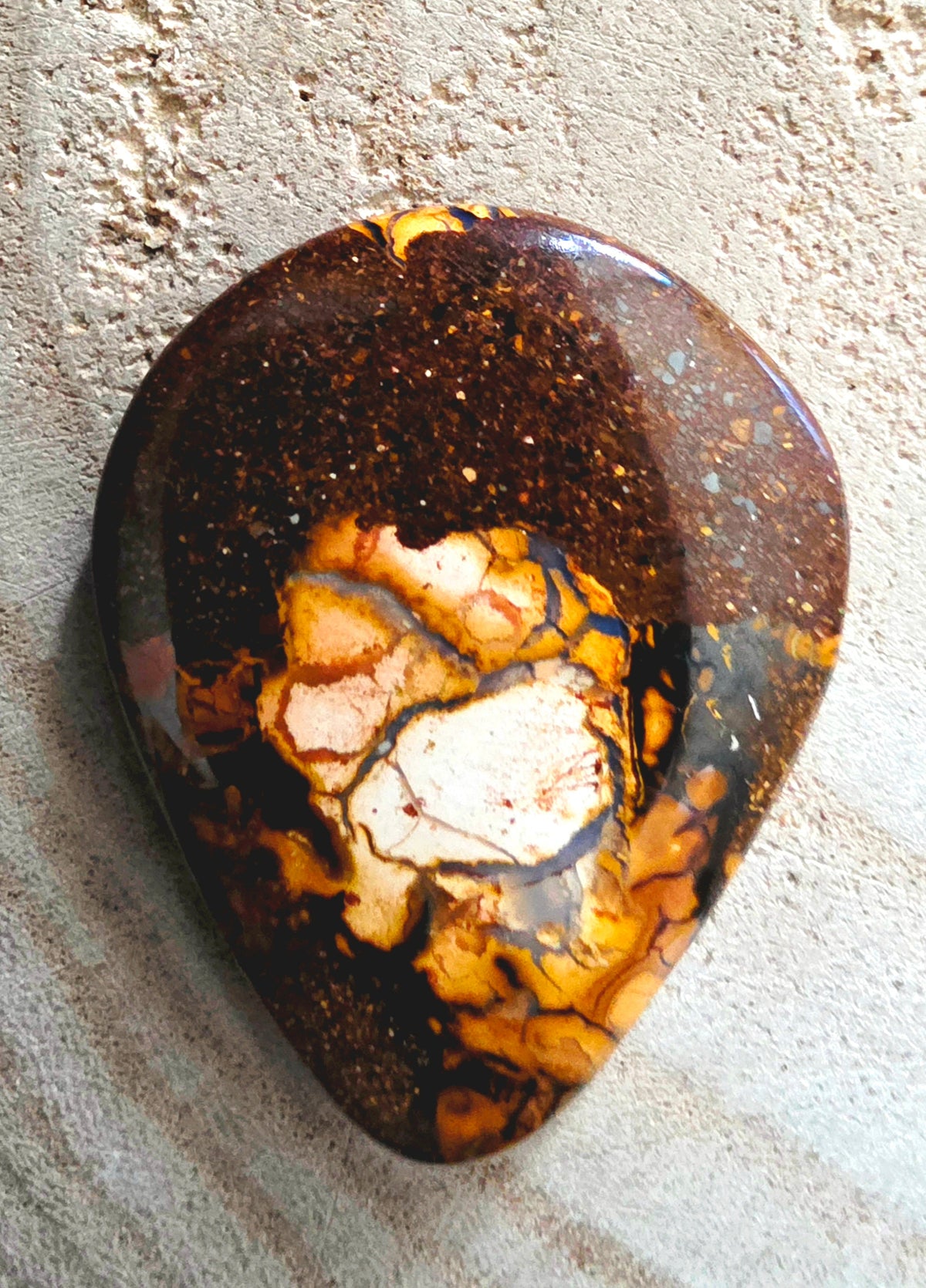A teardrop-shaped polished gemstone cabochon with a marbled pattern of dark brown, golden yellow, and cream, resting on a textured sandy surface.