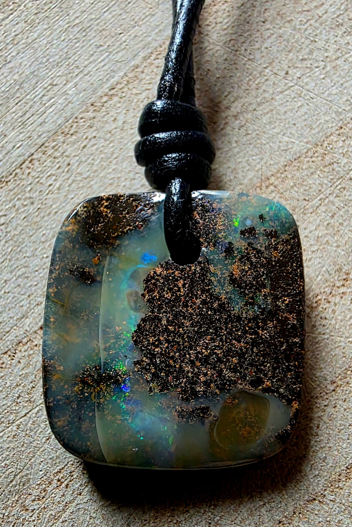 A square opal pendant necklace with a black cord, resting on a textured beige surface. The pendant features a natural opal pattern with dark brown matrix, grey-green base, and flashes of blue and green iridescence.