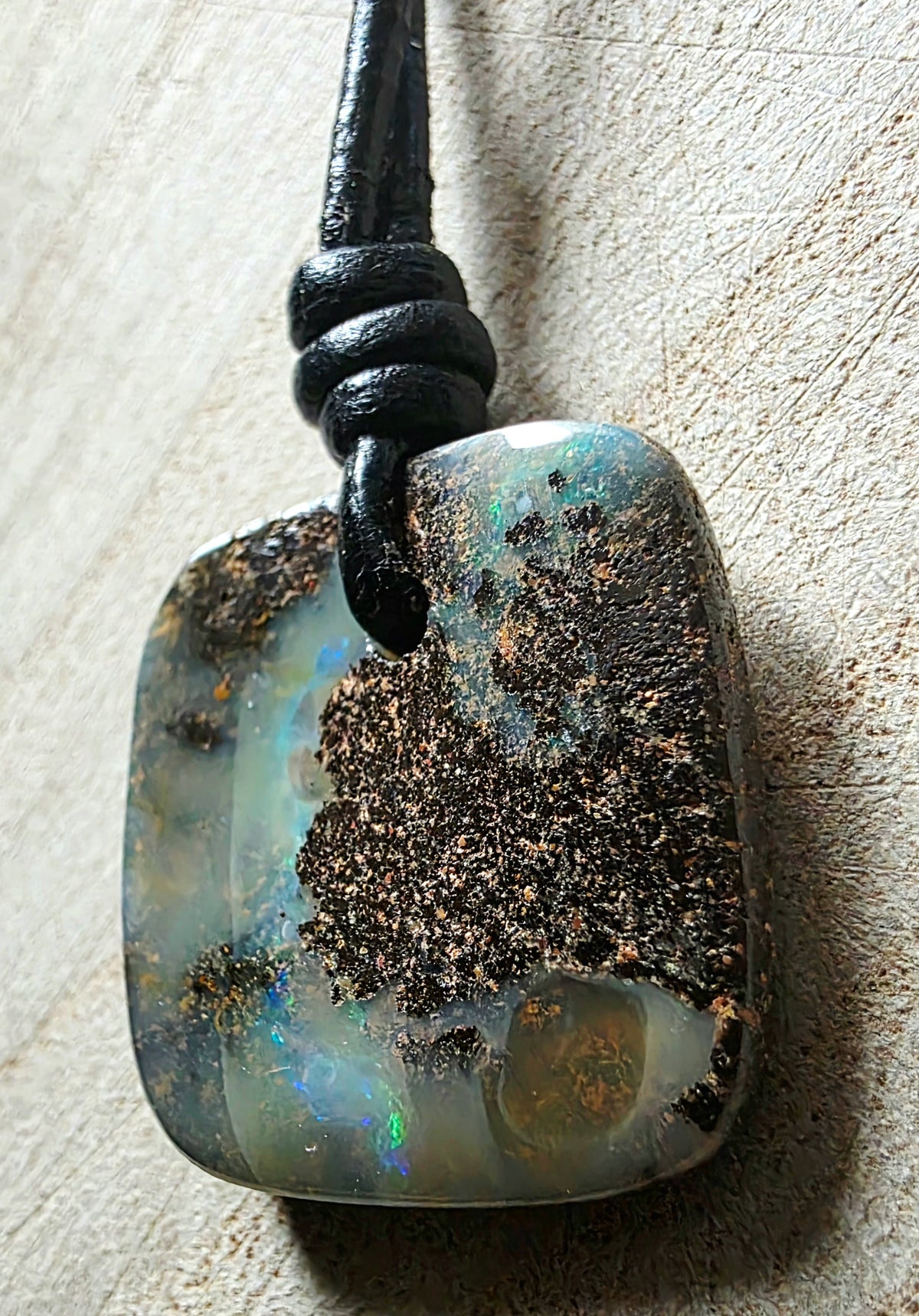 A polished opal pendant with a black cord, resting on a textured beige surface. The pendant features natural opal inclusions and a prominent dark brown matrix, with hints of iridescent blue and green flashes in the opal.