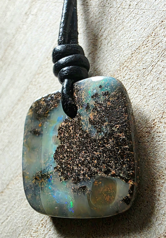 A polished opal pendant with a black cord, resting on a textured beige surface. The pendant features natural opal inclusions and a prominent dark brown matrix, with hints of iridescent blue and green flashes in the opal.