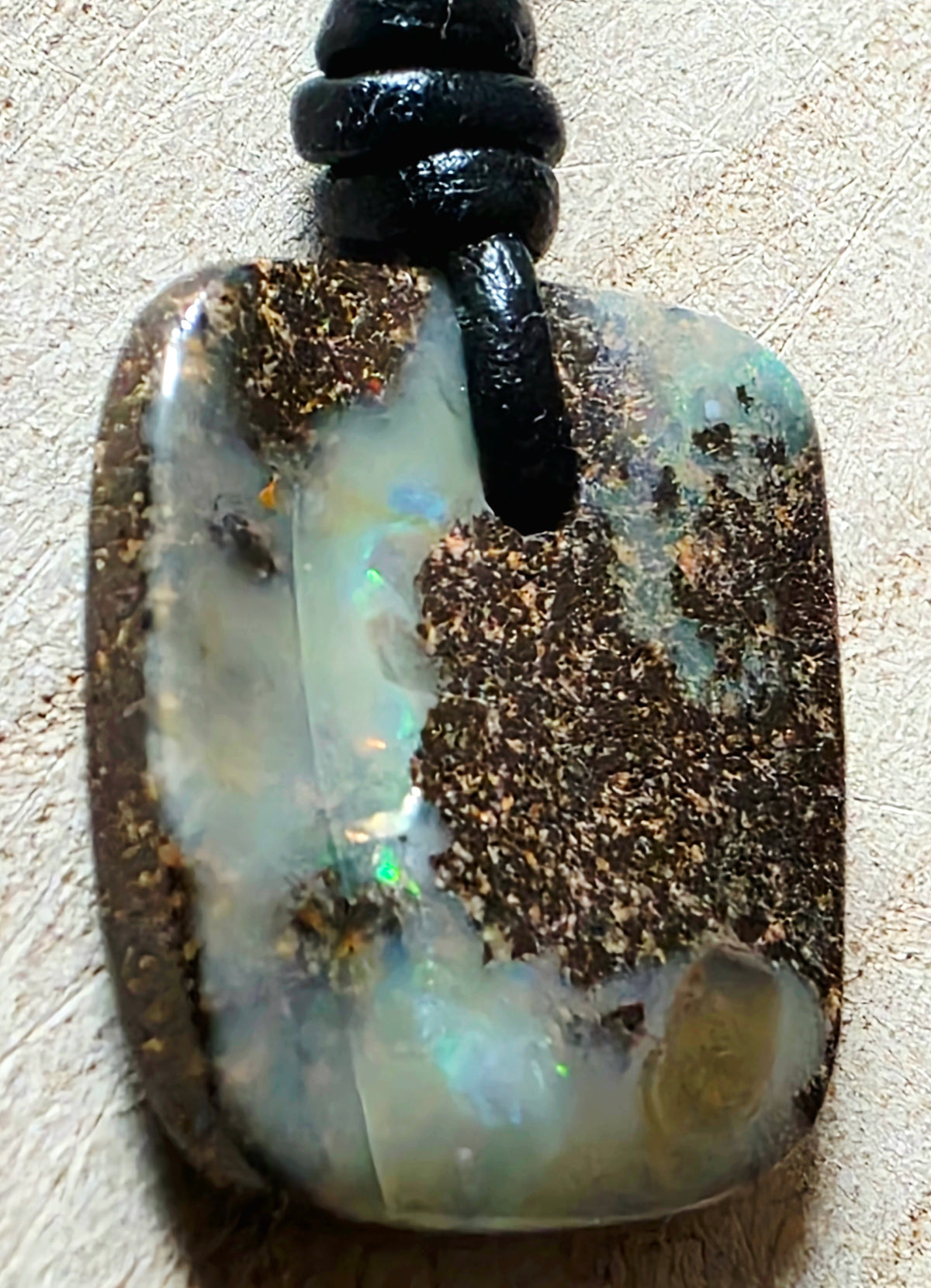 A natural opal pendant with a black cord, featuring a mix of milky white, iridescent blue-green flashes, and prominent dark brown matrix, shown on a textured beige background.