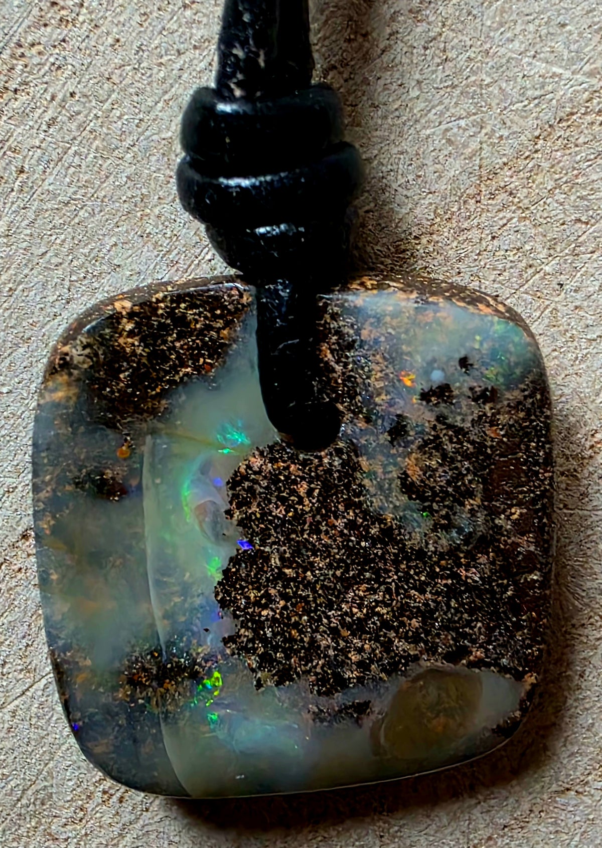 A square opal pendant with a black cord, featuring a mix of iridescent white, blue, and green opal patterns overlaid with dark brown and gold matrix, set against a textured beige background.