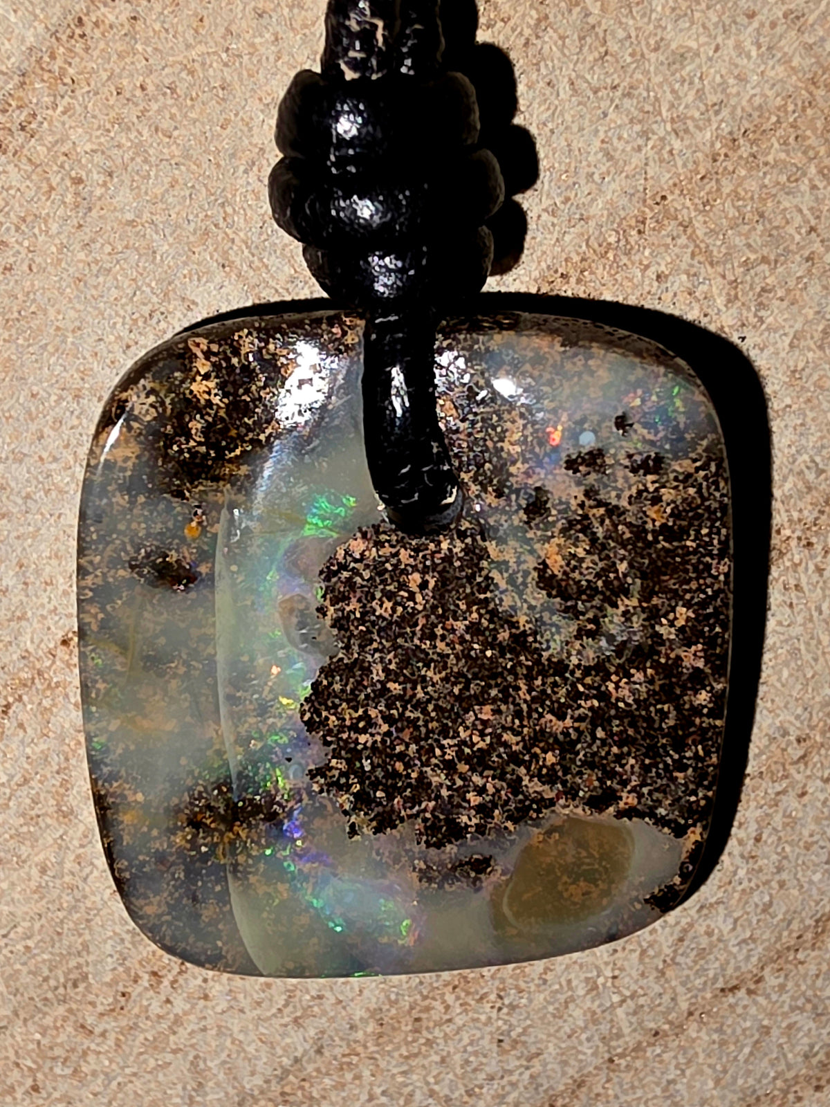 A square opal pendant with rounded corners, featuring a black cord and a background of textured wood. The opal displays a mix of milky white, iridescent flashes of green and blue, and prominent dark brown matrix inclusions.