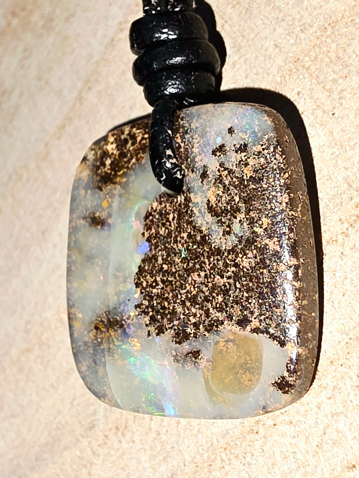 A rectangular cabochon pendant made of opal with dark brown inclusions and a black cord, shown on a beige textured surface.
