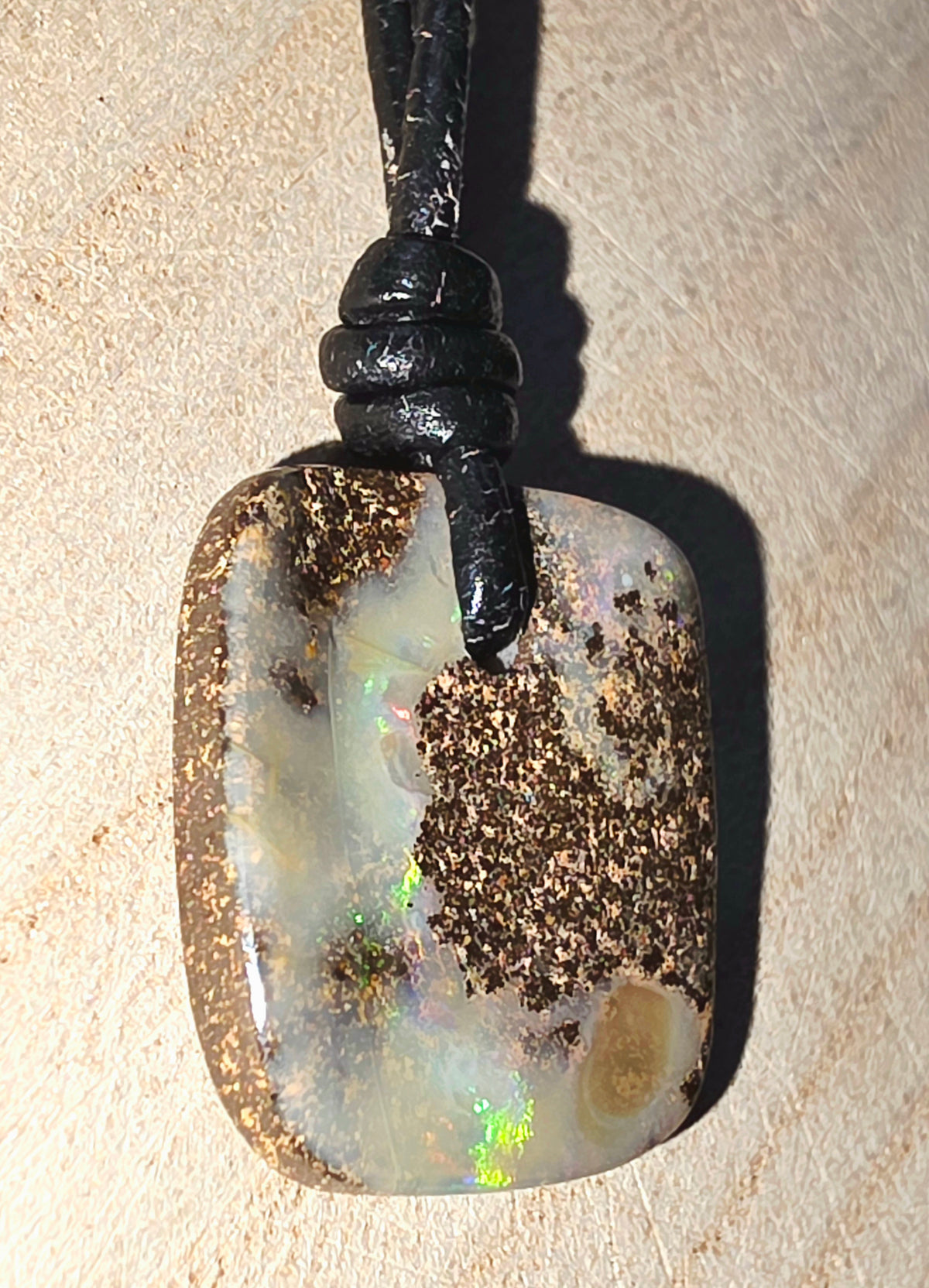 A natural opal pendant with a black cord, displayed on a textured beige surface. The pendant features a mix of white, grey, and brown opal matrix with a vibrant green iridescent flash.