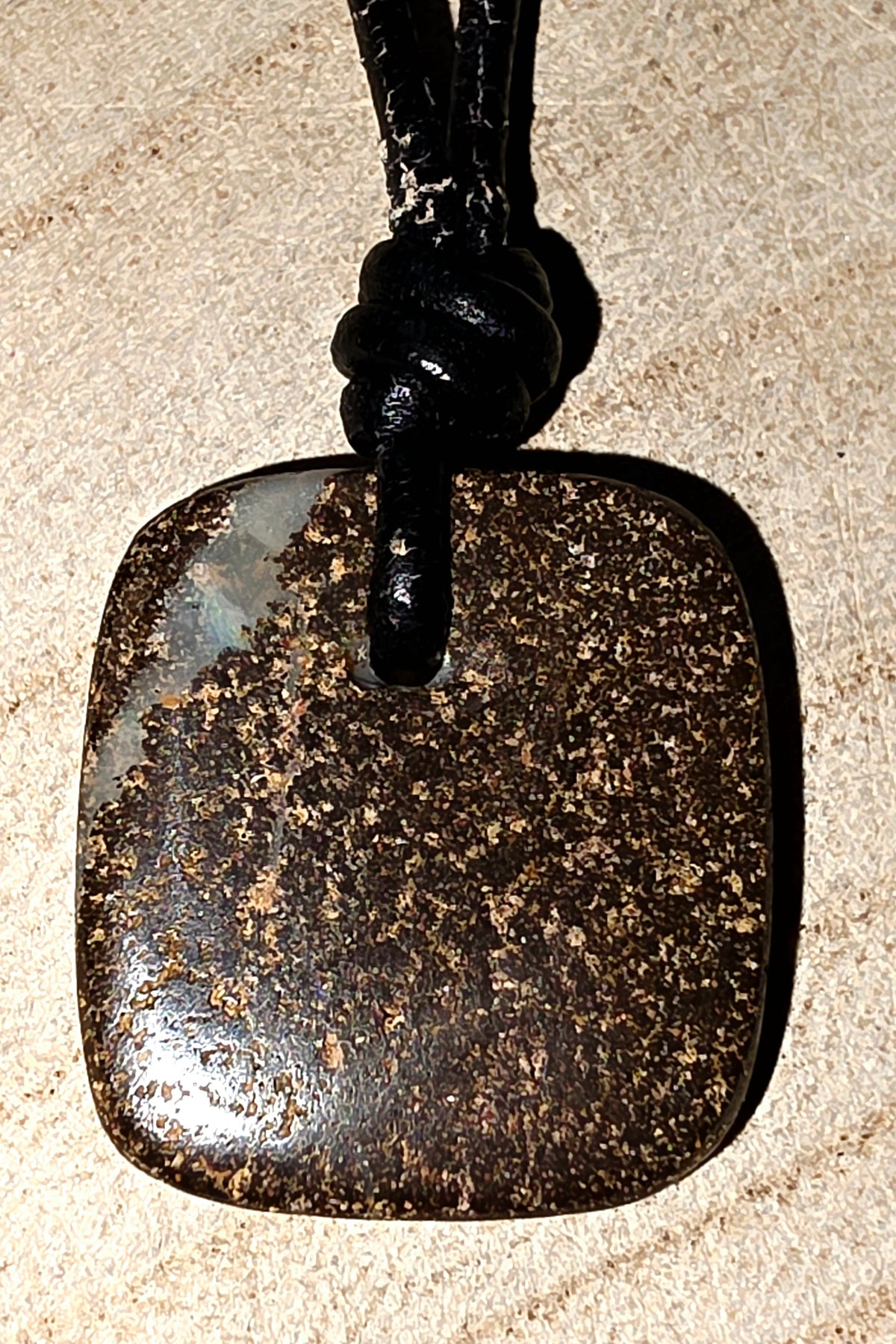 A square dark brown polished stone pendant with natural speckled inclusions, suspended from a black cord on a light beige textured surface.