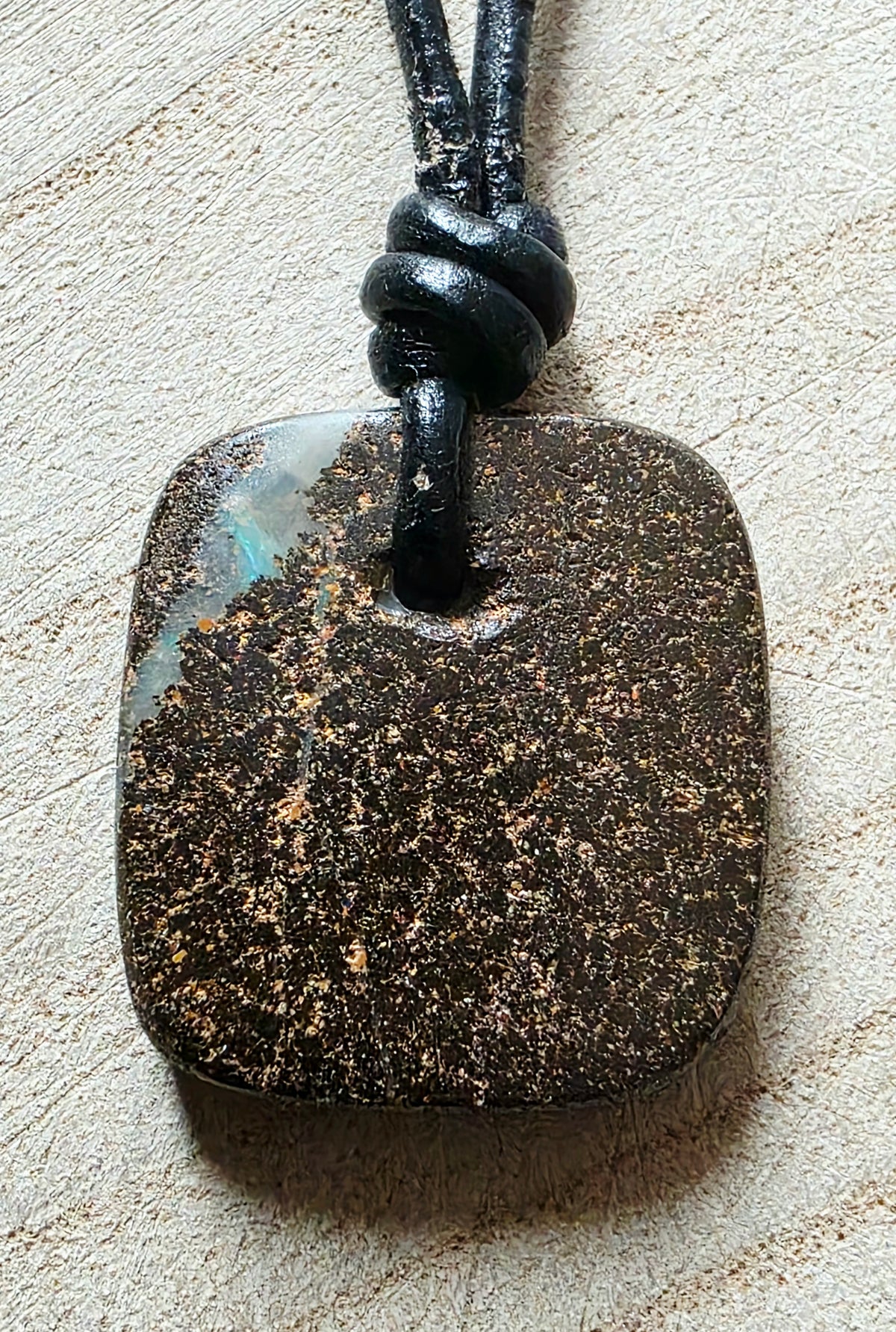 A square dark stone pendant with a natural mottled pattern of brown, black, and subtle iridescent blue-green areas, suspended on a black cord against a light textured background.