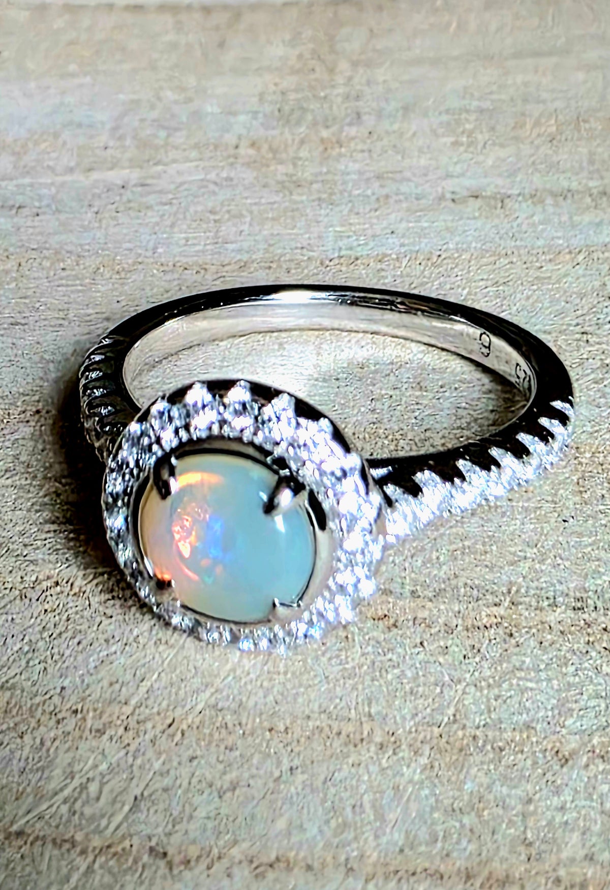 A silver ring featuring a round iridescent opal centre stone surrounded by a halo of small clear stones, with a textured silver band, resting on a textured beige surface.
