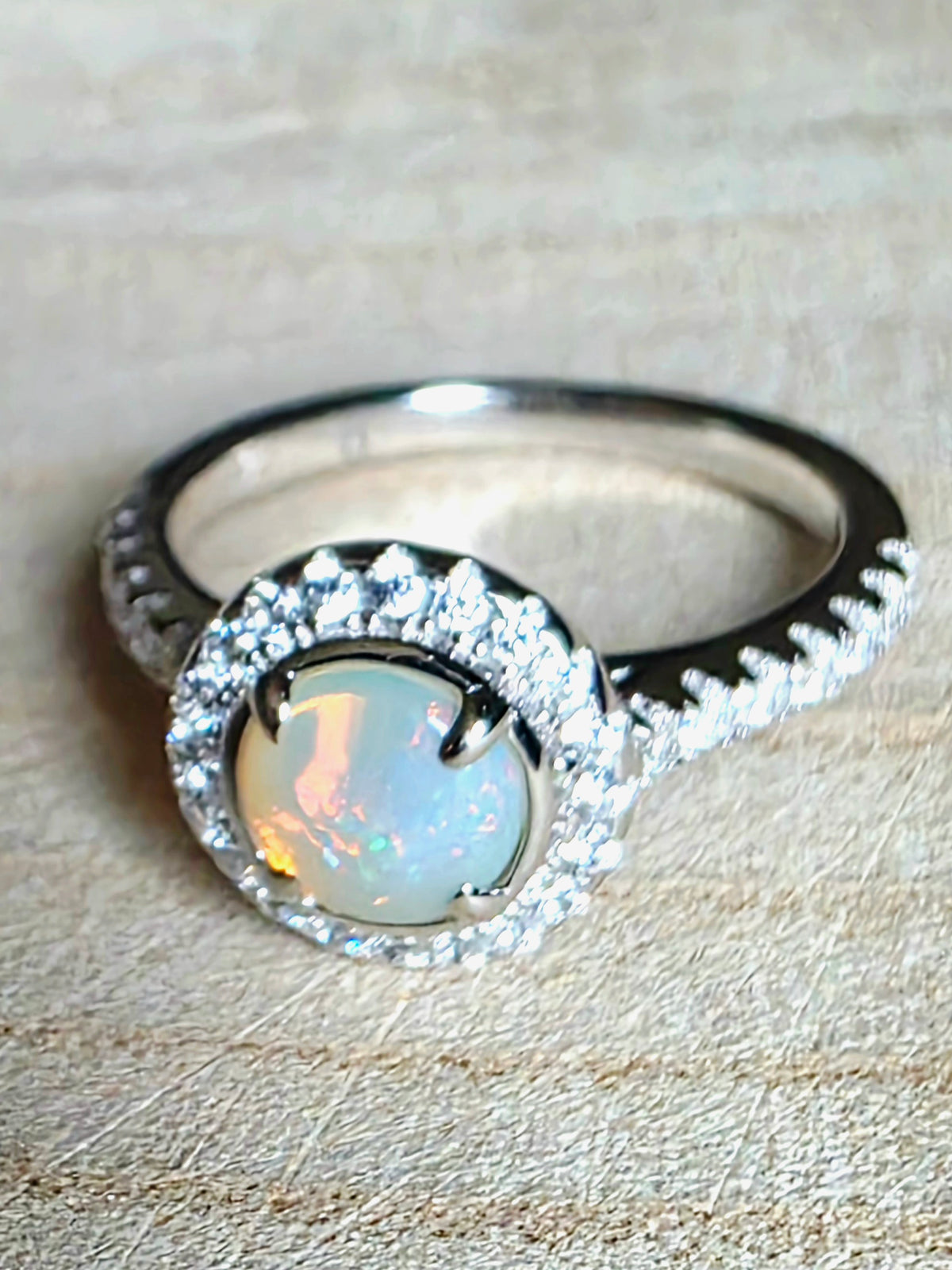 A silver ring featuring a round iridescent white opal centre stone surrounded by a halo of small clear stones, with a textured silver band, resting on a beige textured surface.