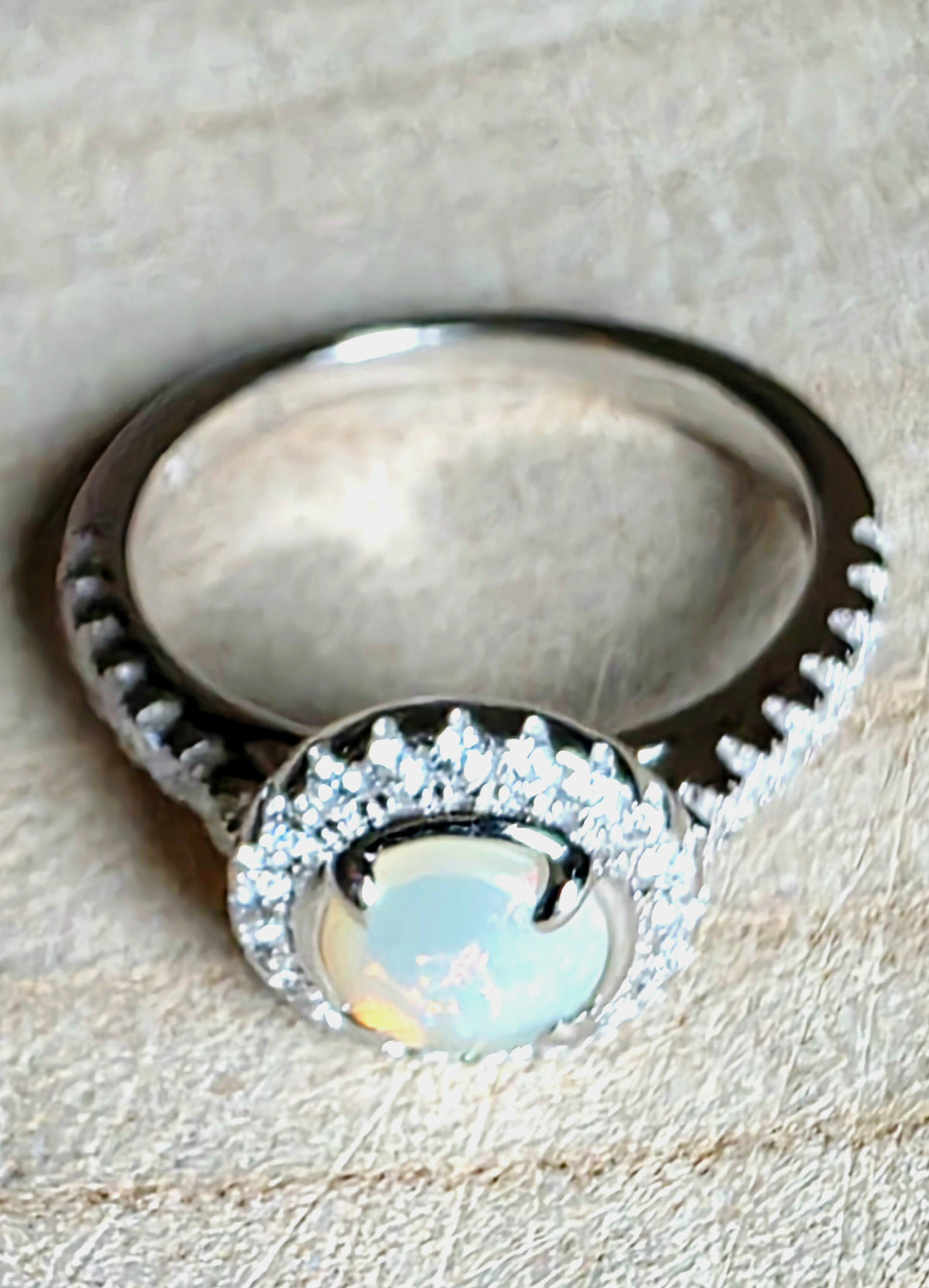 A silver ring featuring a round iridescent white opal centre stone surrounded by a halo of small clear stones, set on a textured beige surface.