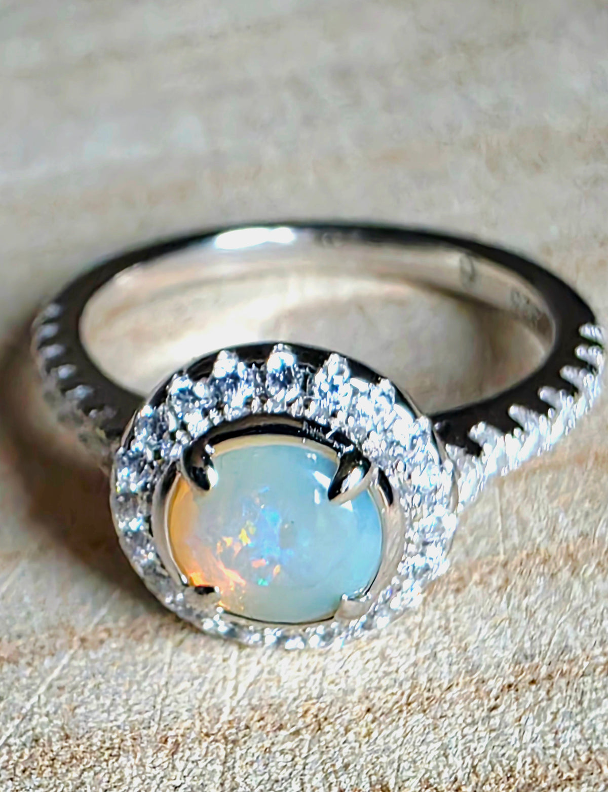 A silver ring featuring a round iridescent crystal opal centre stone surrounded by a halo of small clear stones, set on a textured beige surface
