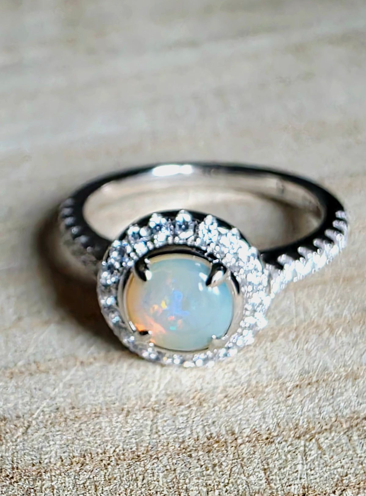 A silver ring featuring a round white opal centre stone surrounded by a halo of small clear stones, set on a textured beige fabric background.