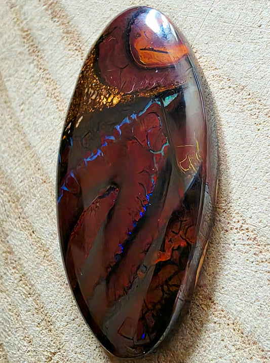 A polished oval opal gemstone with a dark reddish-brown base and vivid iridescent blue, orange, and gold flashes, resting on a textured beige fabric background.