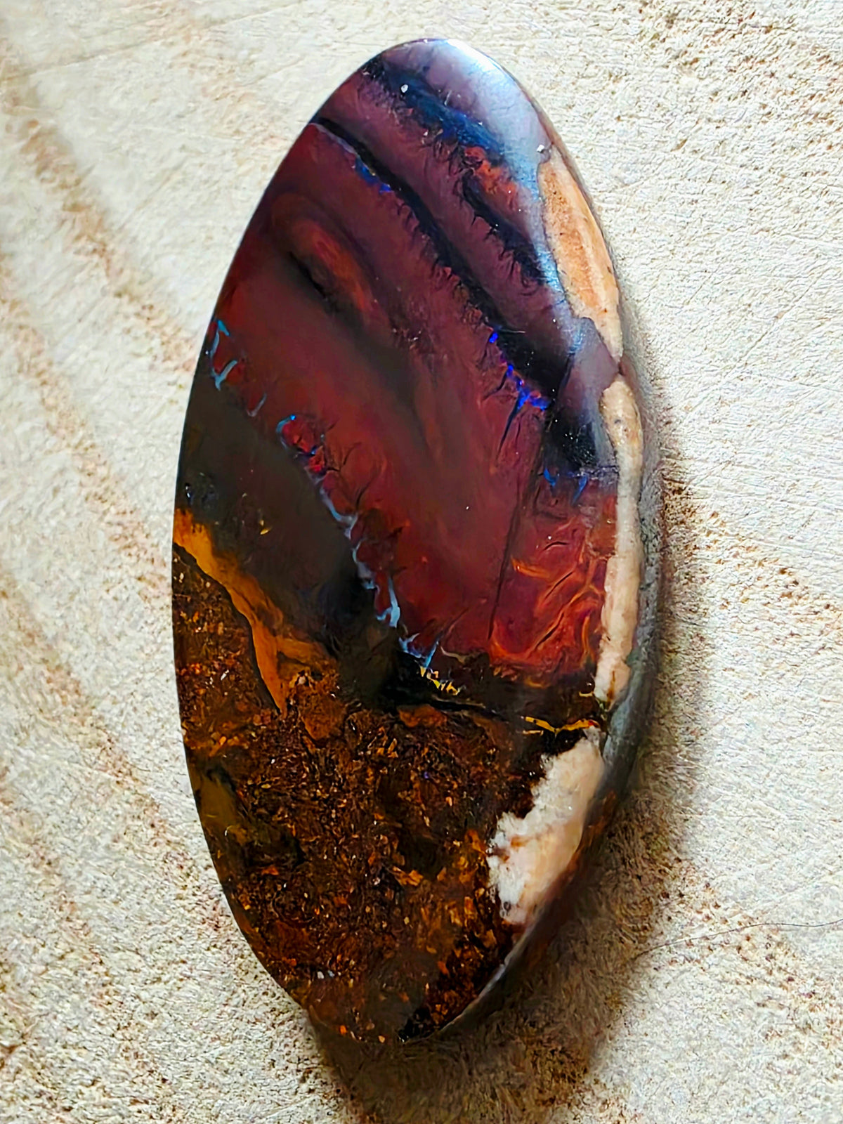 A polished oval gemstone with a marbled pattern of deep red, black, brown, and cream, resting on a textured beige fabric background.