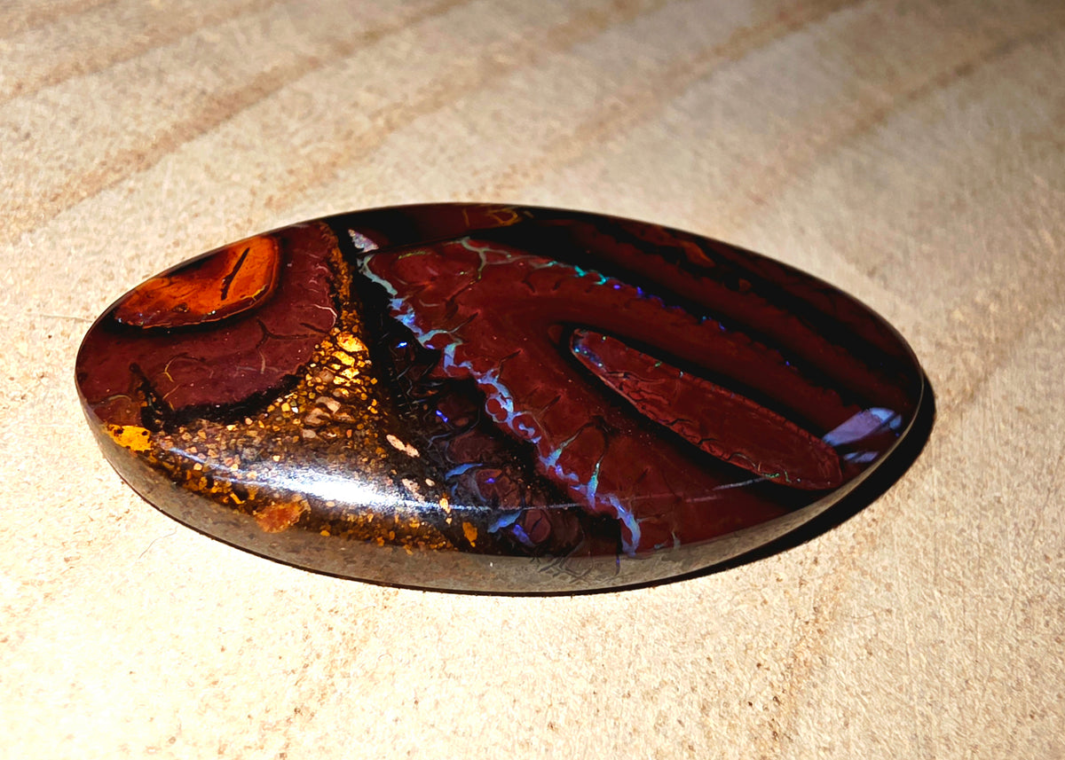 A polished oval cabochon gemstone featuring a rich marbled pattern of deep burgundy, black, gold, and subtle iridescent blue-purple veins, resting on a textured beige sand surface.