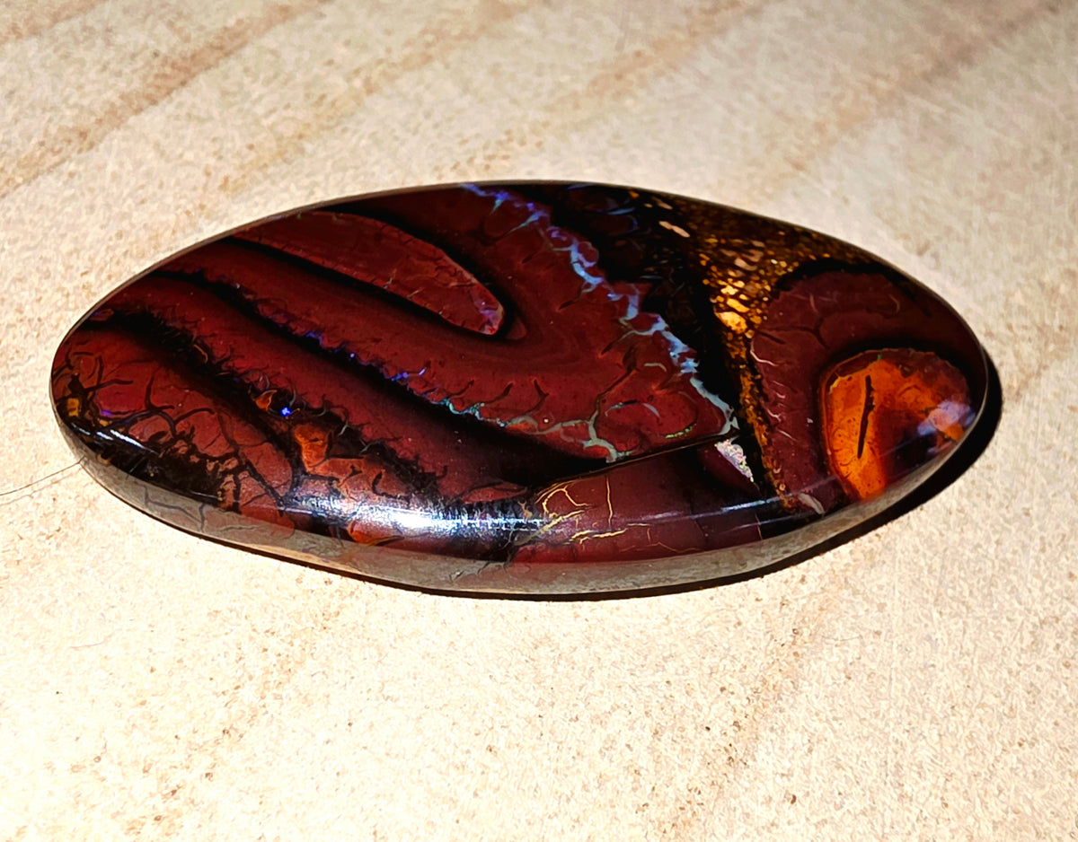 A polished oval cabochon gemstone featuring a marbled pattern of deep red, black, and orange with subtle blue iridescence, resting on a textured beige sand surface.