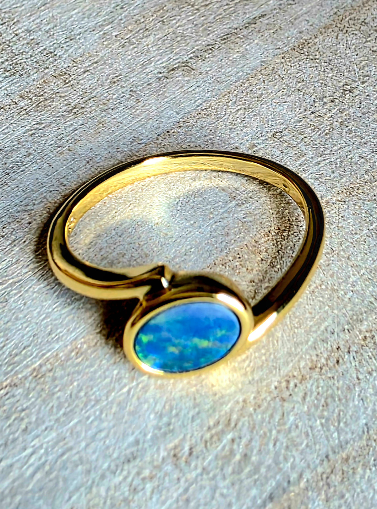 A gold ring with a round, vibrant blue opal gemstone, resting on a textured light grey surface.