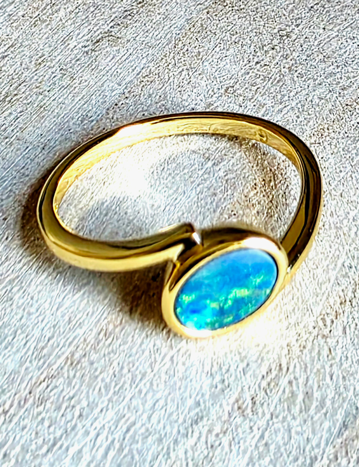 A gold-plated open band ring featuring a bezel-set oval blue opal gemstone, resting on a textured light grey surface.