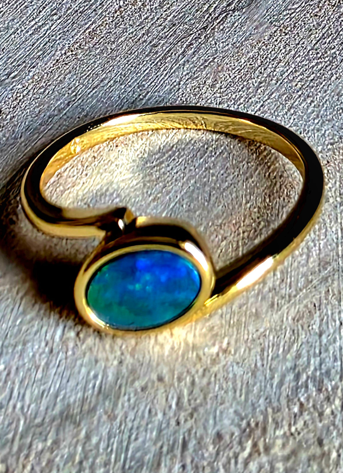 A gold ring with a bezel-set oval blue opal stone, photographed close-up on a textured grey surface.