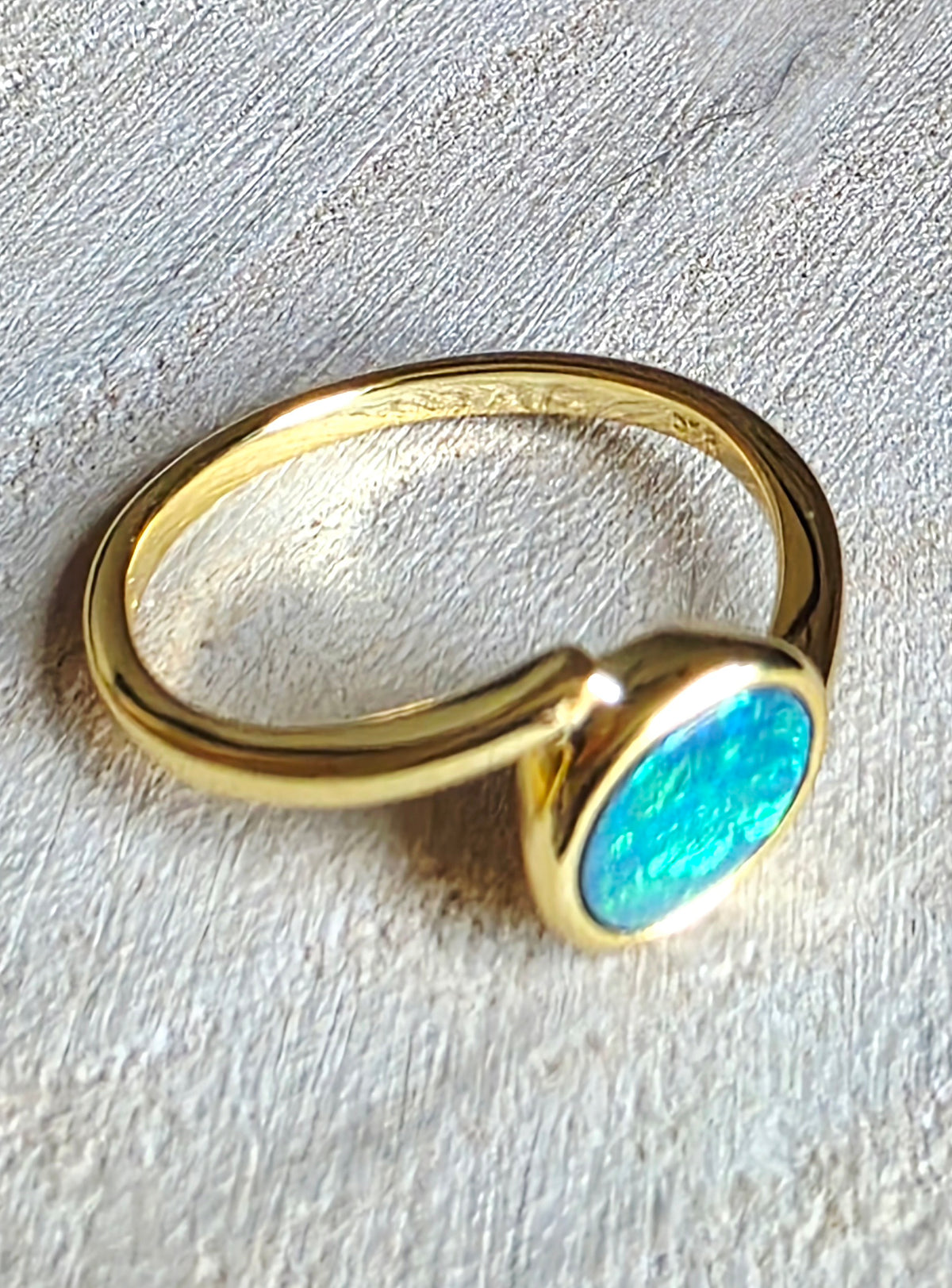 A gold-plated ring with a curved band and a bezel-set oval turquoise stone, resting on a textured light grey surface.
