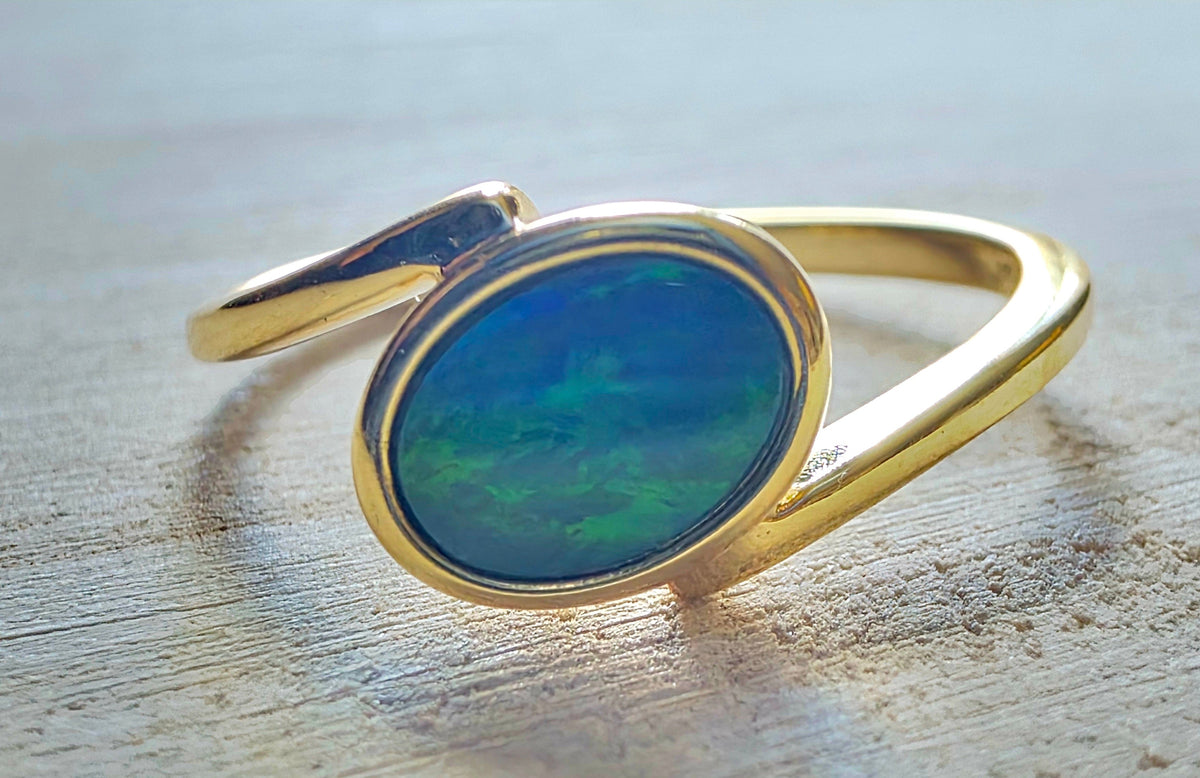 A gold ring with an oval-shaped blue-green iridescent opal gemstone, resting on a light-coloured wooden surface.