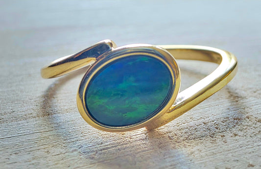 A gold ring with an oval-shaped blue-green iridescent opal gemstone, resting on a light-coloured wooden surface.