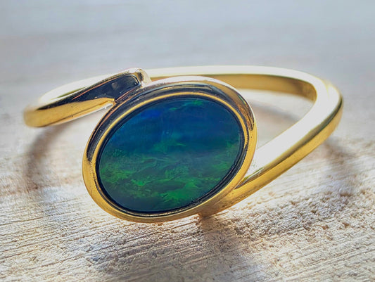 A gold ring with an oval cabochon opal gemstone showing vibrant green and blue play-of-colour, resting on a textured light wood surface.