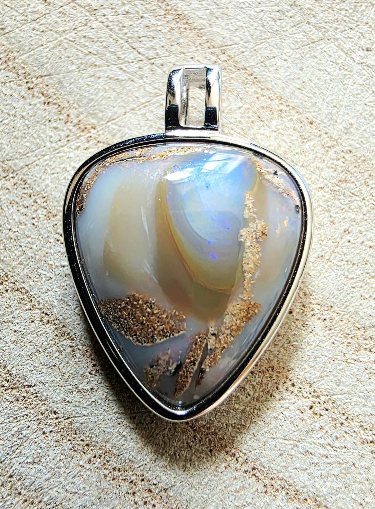 A triangular pendant featuring a polished opal-like stone with iridescent white, blue, and cream tones, set in a silver metal bezel with a bail, photographed on a textured beige surface.