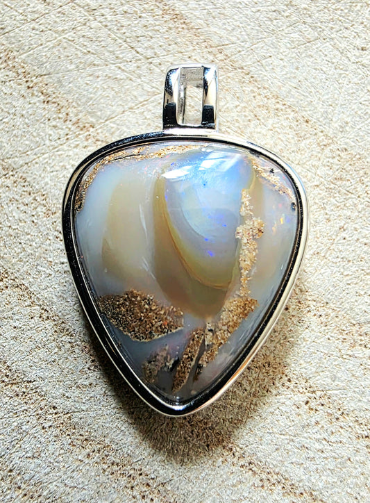 A triangular pendant featuring a polished opal-like stone with iridescent white, blue, and cream tones, set in a silver metal bezel with a bail, photographed on a textured beige surface.
