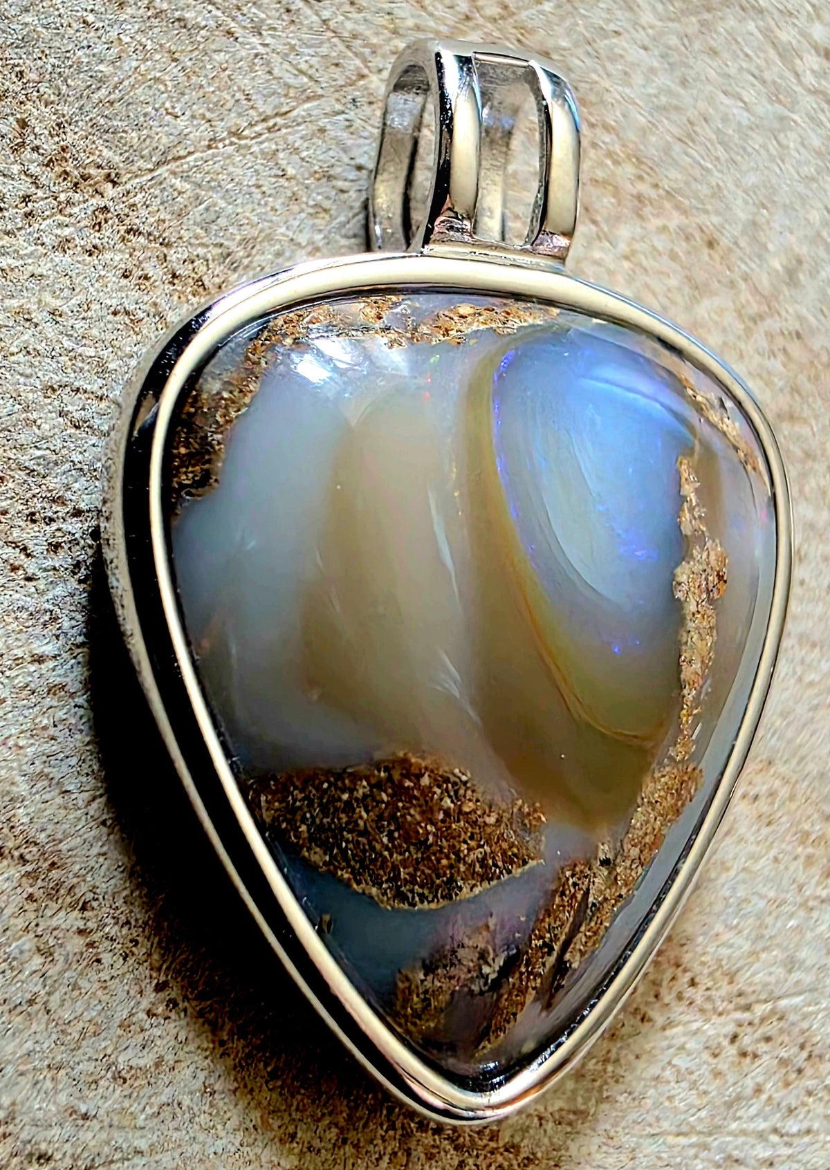 A triangular pendant featuring a polished iridescent stone with blue, grey, and brown bands, set in a polished silver-tone metal bezel with a bail, resting on a textured beige surface.