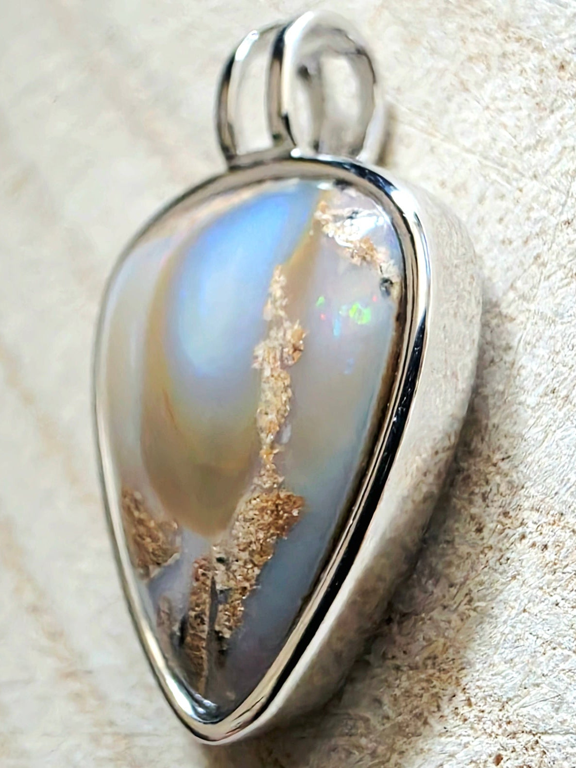 A silver teardrop-shaped pendant featuring a polished iridescent stone with blue, green, and gold inclusions, resting on a textured beige fabric background.