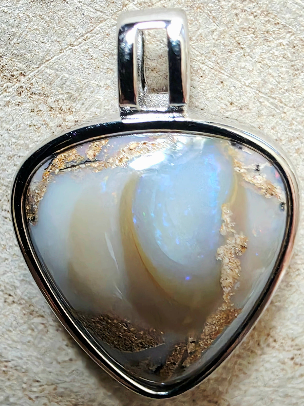 A triangular pendant featuring a white and iridescent opal stone with gold inclusions, set in a polished silver metal bezel with a hanging loop.