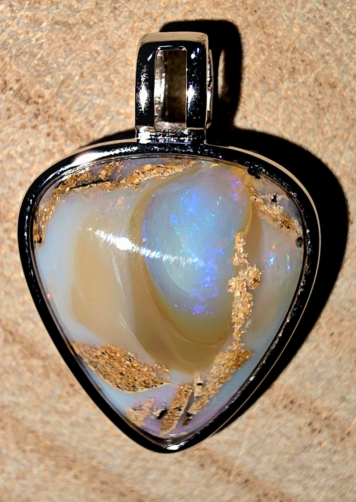 A heart-shaped opal pendant with a silver metal border and bail, resting on a textured beige surface. The opal features a prominent iridescent blue-white centre with golden crystalline inclusions.
