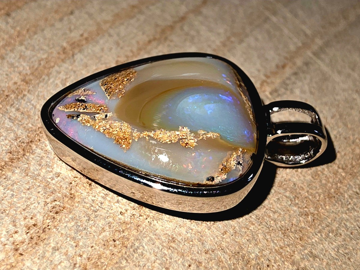 A heart pendant featuring a polished iridescent opal stone with gold inclusions, set in a polished silver-tone bezel with a loop bail, resting on a textured beige stone surface.