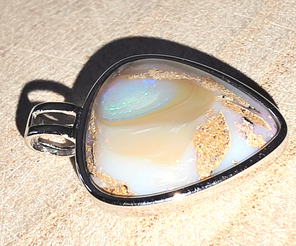 A heart pendant featuring a white and iridescent opal inlay with gold flecks, set in a polished silver-tone bezel with a bail, resting on a textured beige surface.