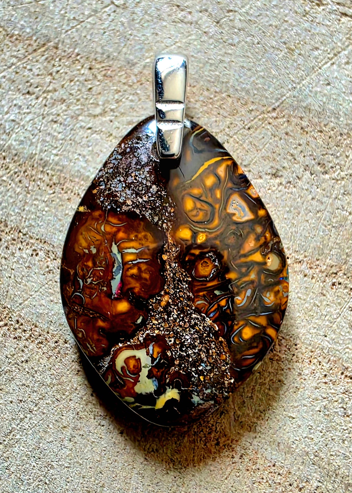 A teardrop-shaped pendant featuring a marbled pattern of brown, amber, black, and cream swirls, accented with a textured dark brown top section and a silver bail, resting on a textured beige surface.