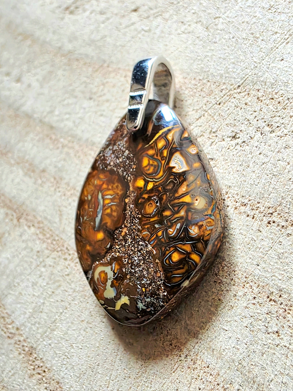 A teardrop-shaped pendant featuring a highly patterned brown and gold banded stone, suspended from a polished silver bail, set against a textured beige background.