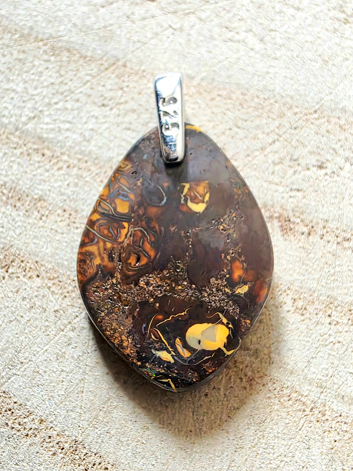 A teardrop-shaped pendant featuring a polished dark brown and black pattern with prominent golden-yellow inclusions, suspended from a silver bail engraved with '925'.