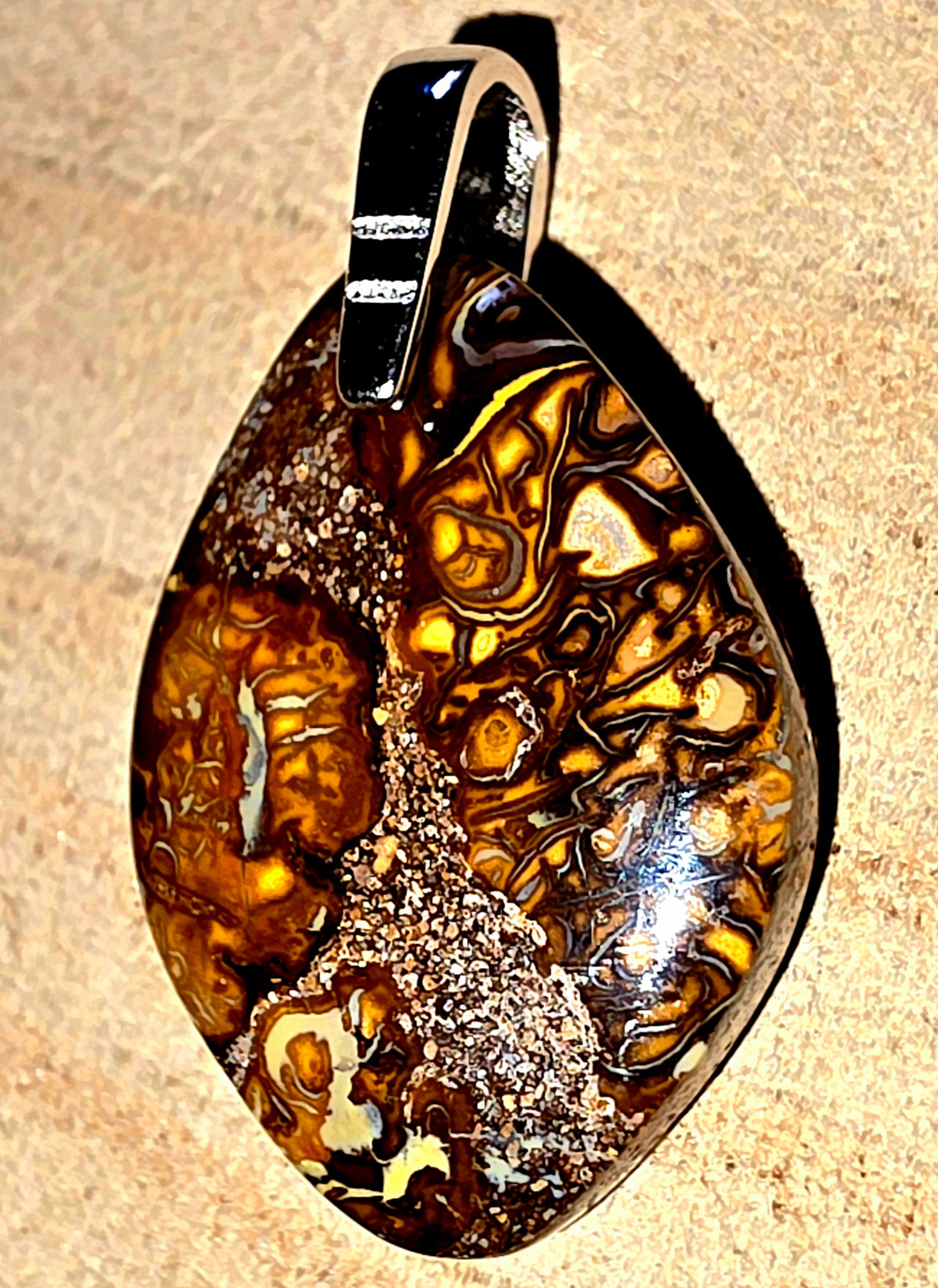 A teardrop-shaped pendant featuring a polished brown and amber marbled pattern with a 925 silver bail, displayed on a textured beige background.