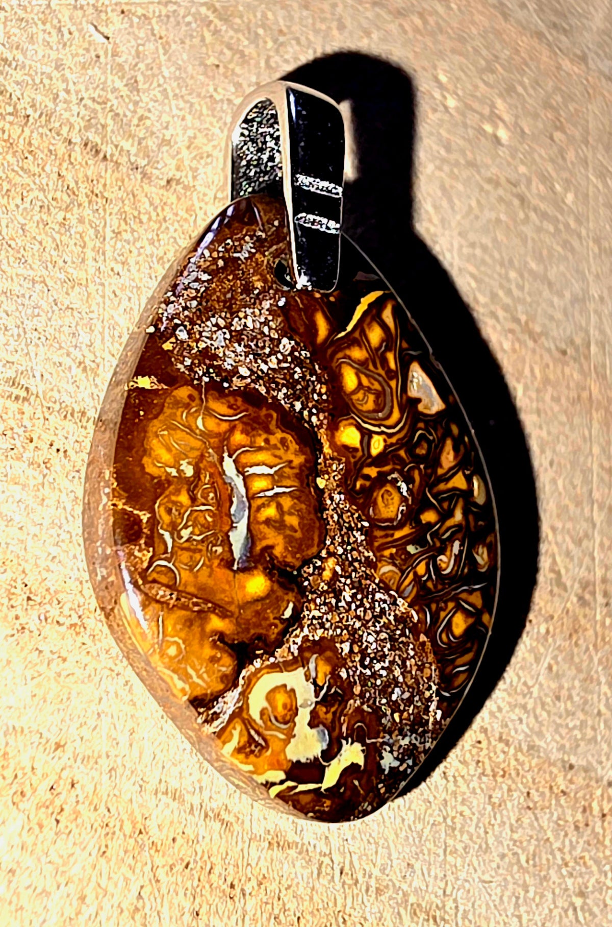 A teardrop-shaped pendant featuring a polished brown and amber marbled opal stone with a silver bail, resting on a textured beige surface.