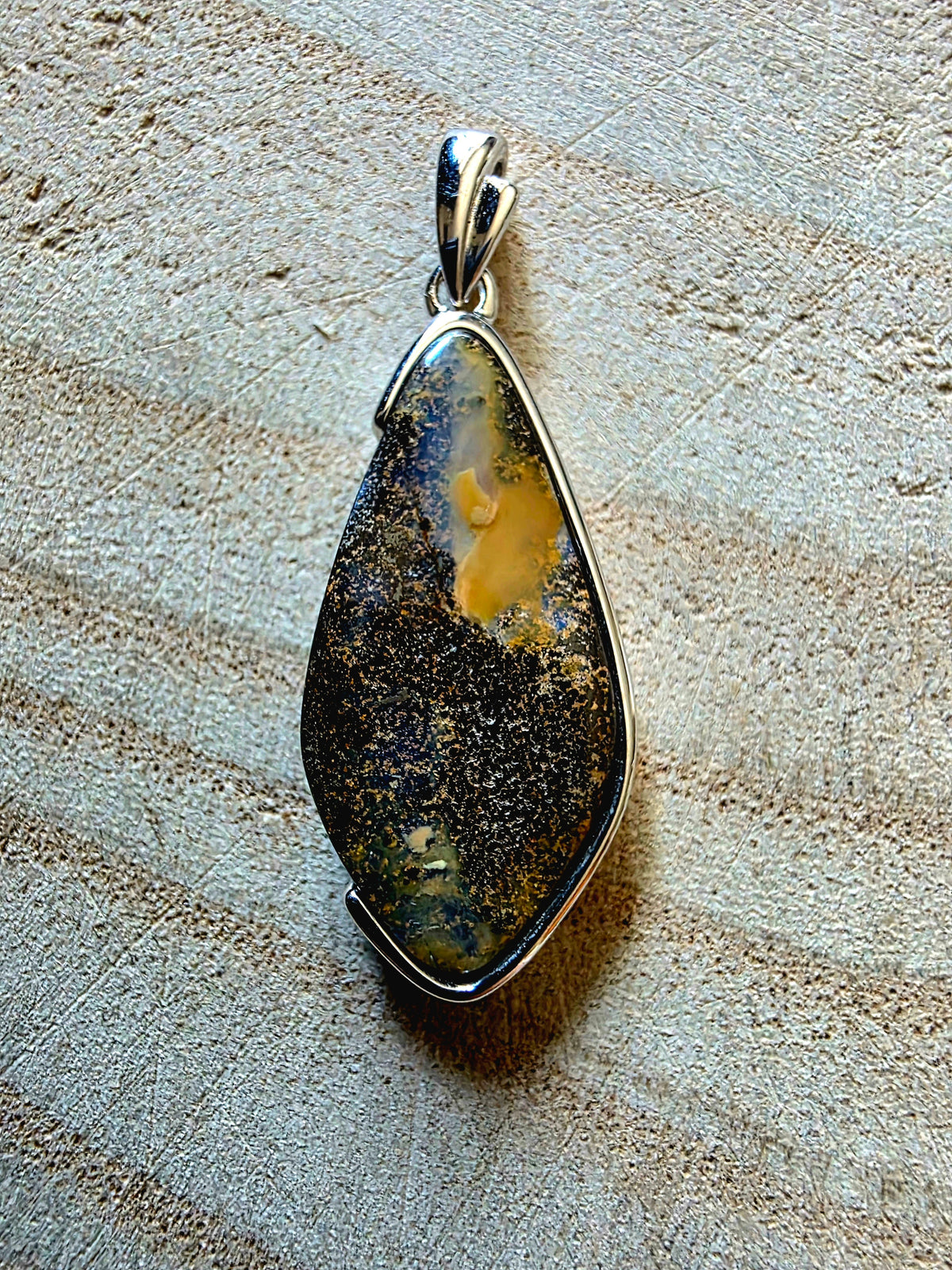 A teardrop-shaped pendant featuring a natural opal dark brown and black stone with a prominent yellow inlay, set in a polished silver bezel, displayed on a textured beige surface.