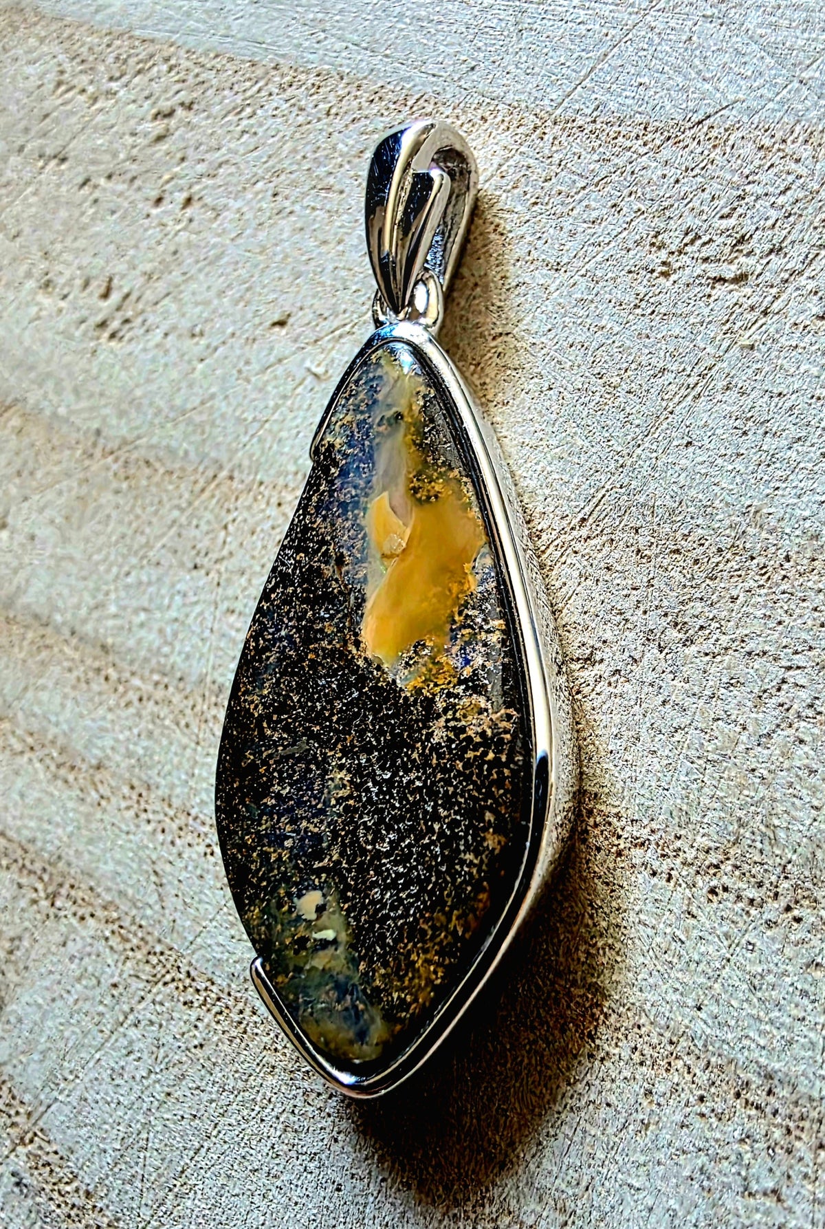 A teardrop-shaped pendant featuring a polished silver bezel set with a natural opal stone that displays a mix of black, brown, yellow, and grey crystalline patterns, resting on a textured beige stone surface.